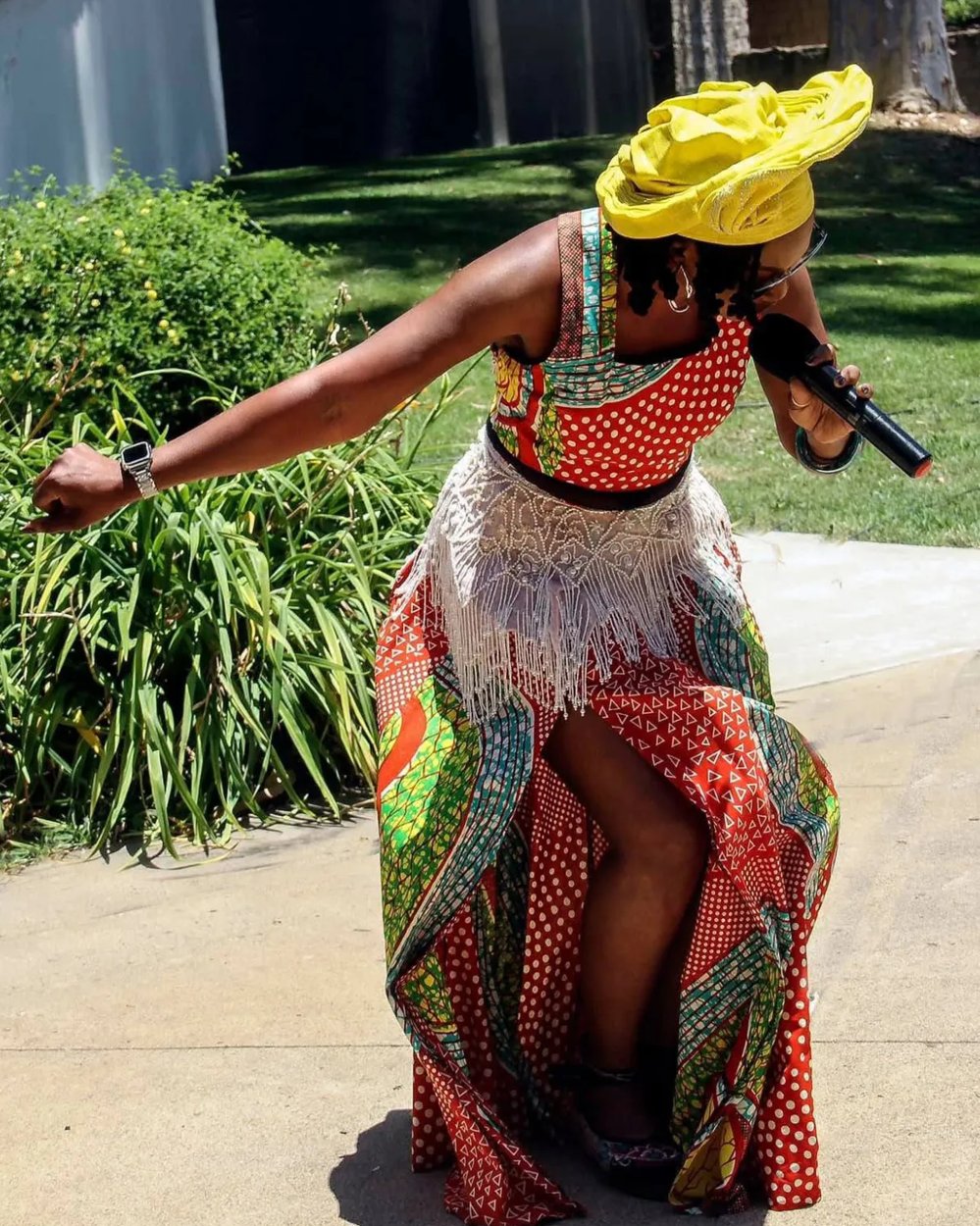 Buki Domingos in a yellow gele and a red polka-dot African print dress, leaning into a song with a microphone